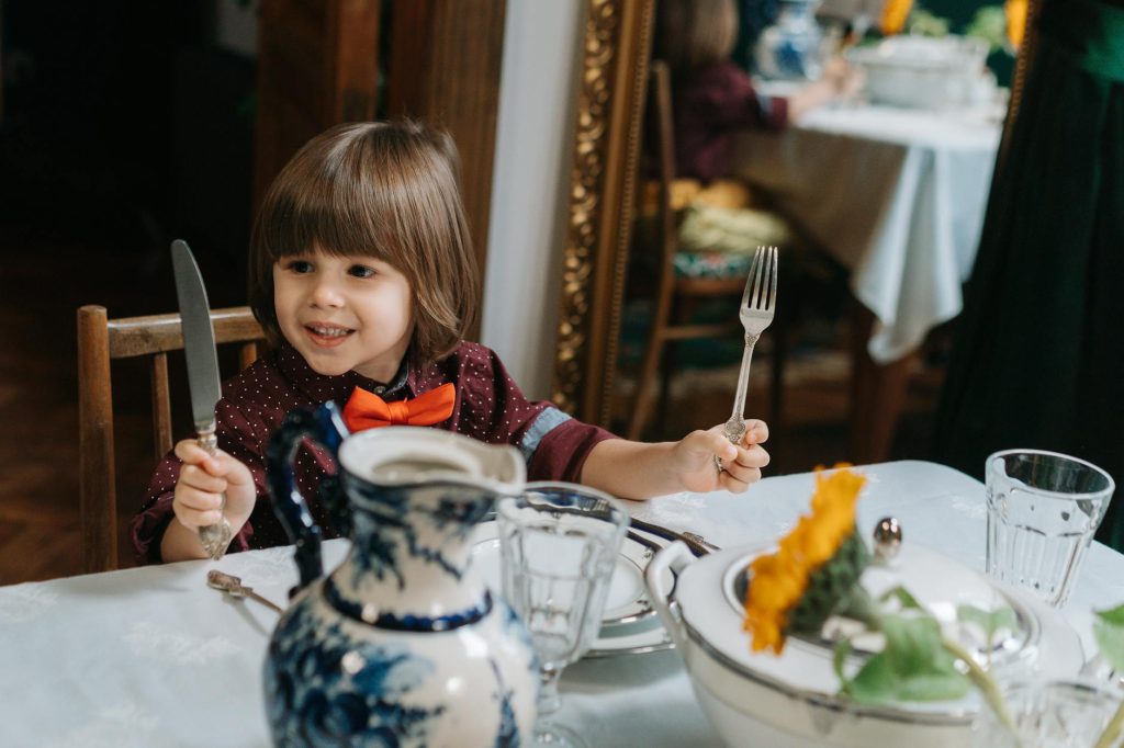 a cute little boy sitting on a chair while holding fork and knife